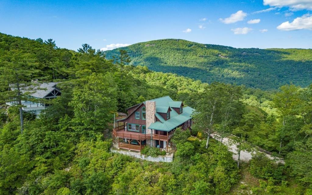 Une grande cabane en bois à plusieurs étages, surmontée d’un toit végétalisé, entourée d’une forêt dense et de collines.