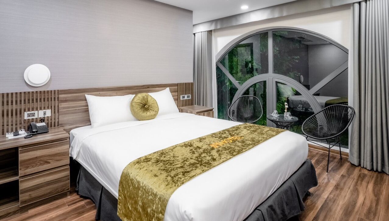 A modern hotel room with a large bed, a wooden headboard, a desk with a phone, and a view of greenery through a large window.