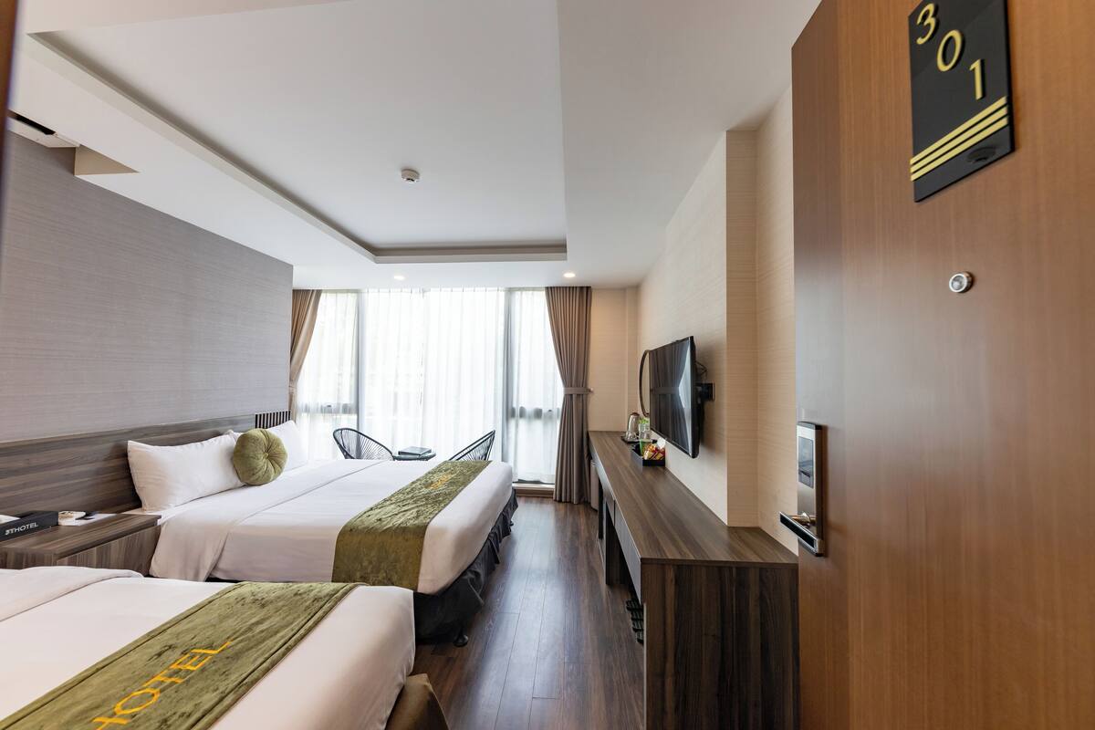 A hotel room with two beds, a TV, a desk, and a large window with curtains.