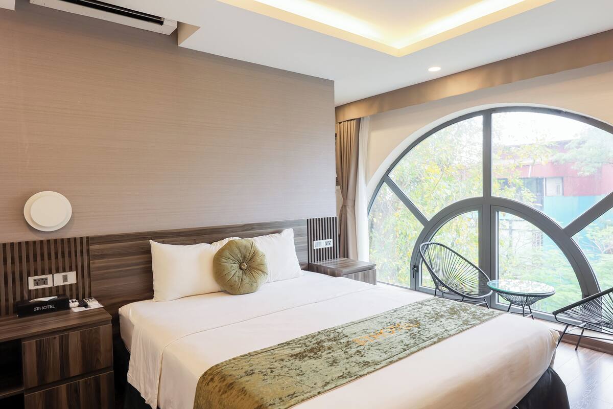 A modern hotel room with a large bed, a small table, two chairs, a nightstand, and a large window with a view of greenery.