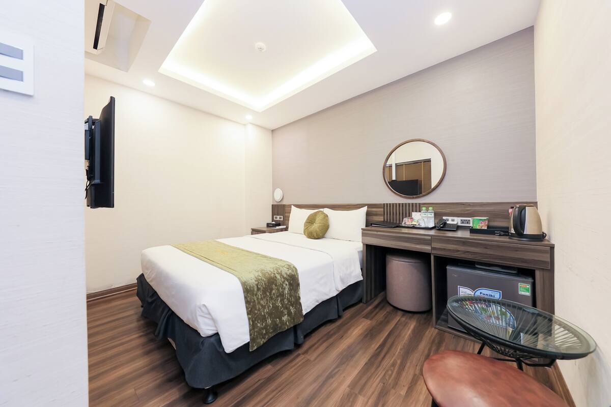 A modern hotel room with a neatly made bed, a desk with a computer, a round mirror, and a small seating area.