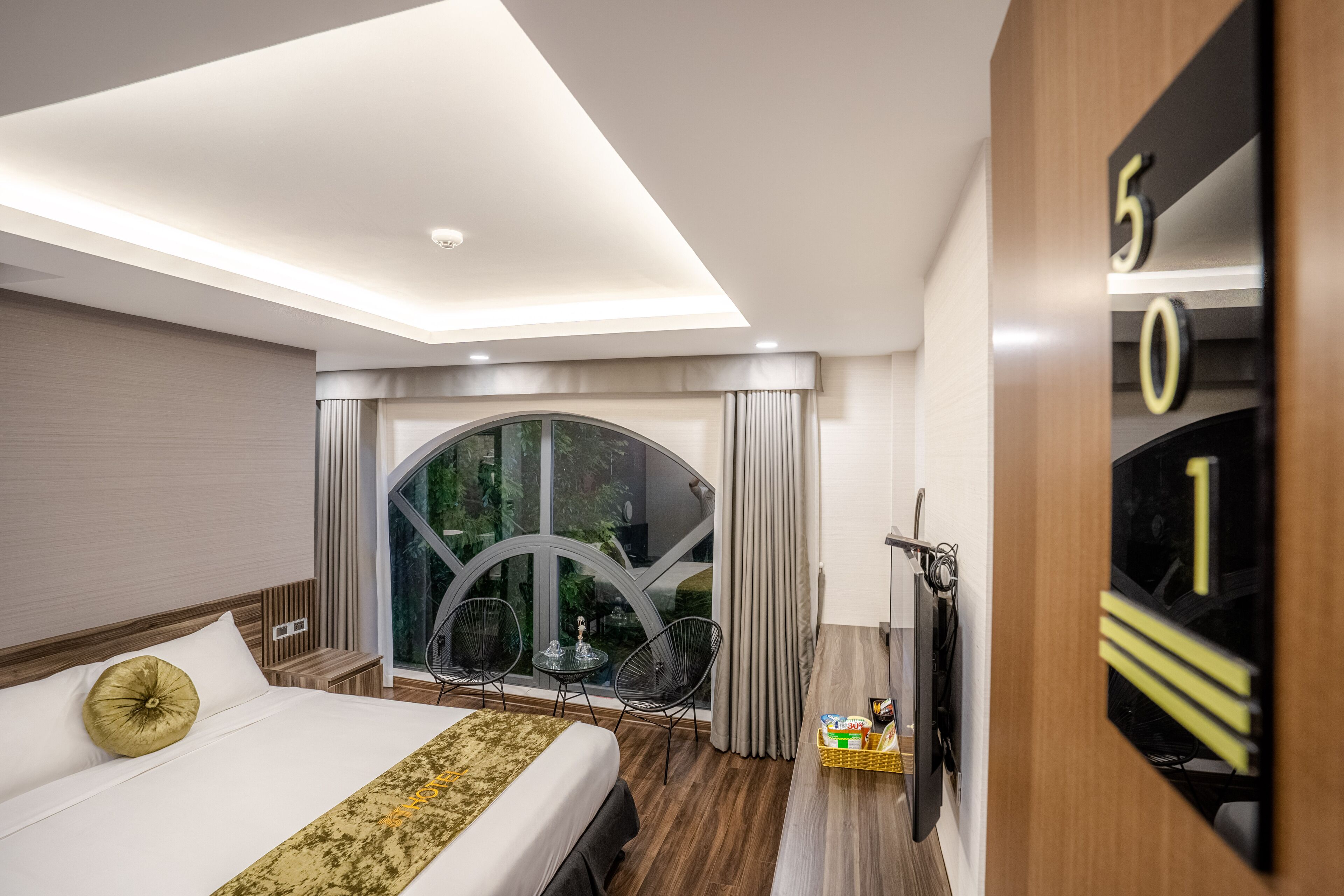 Une chambre d’hôtel moderne, dotée d’un grand lit, d’une petite table, de deux chaises, d’une télévision et d’une grande fenêtre avec des rideaux.