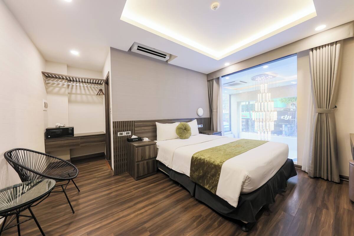A modern hotel room with a large bed, a sitting area with a chair, a bedside table, and a view of the city through a large window.