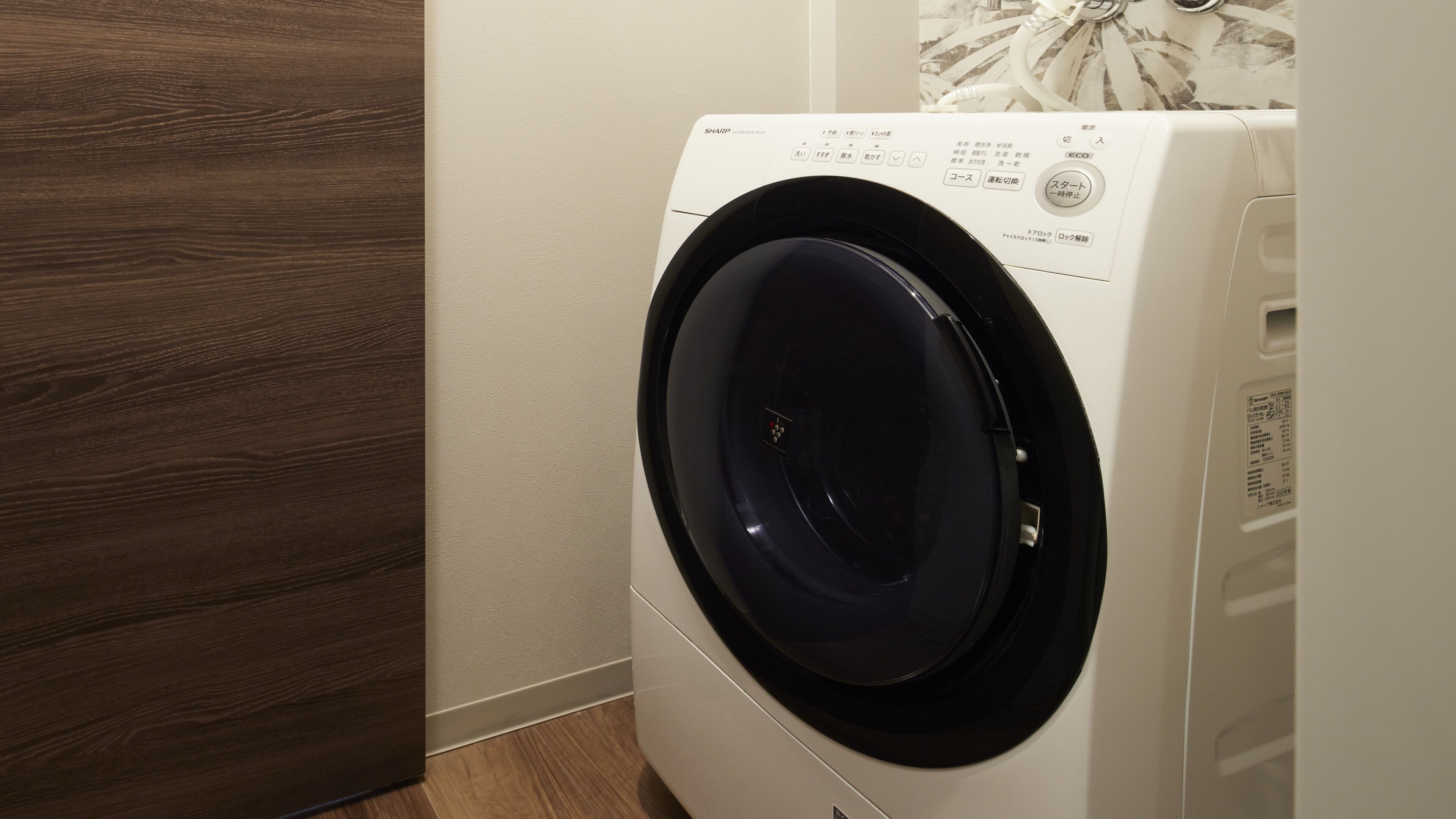 A front view of a white washing machine with a black door, located in a room with wooden flooring and a white wall.