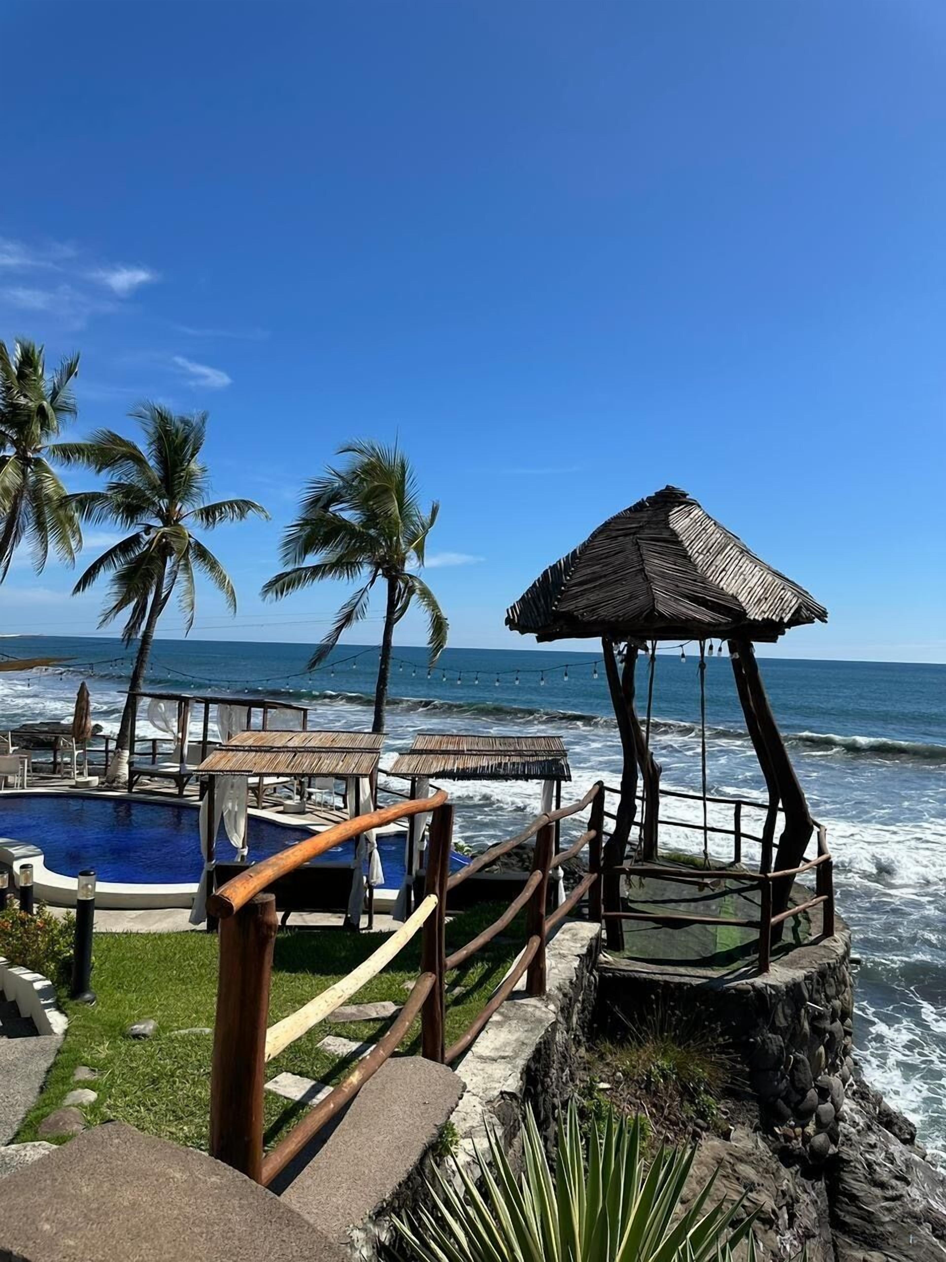 A pool area with a wooden deck, a thatched-roof gazebo, and palm trees by the ocean.