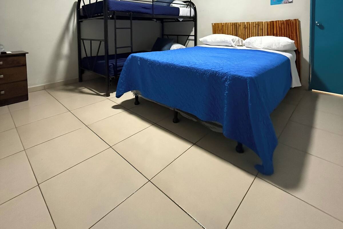 A single bedroom with a bunk bed, a bed with a blue bedspread, a wooden headboard, a dresser, a door, and a wall-mounted picture.