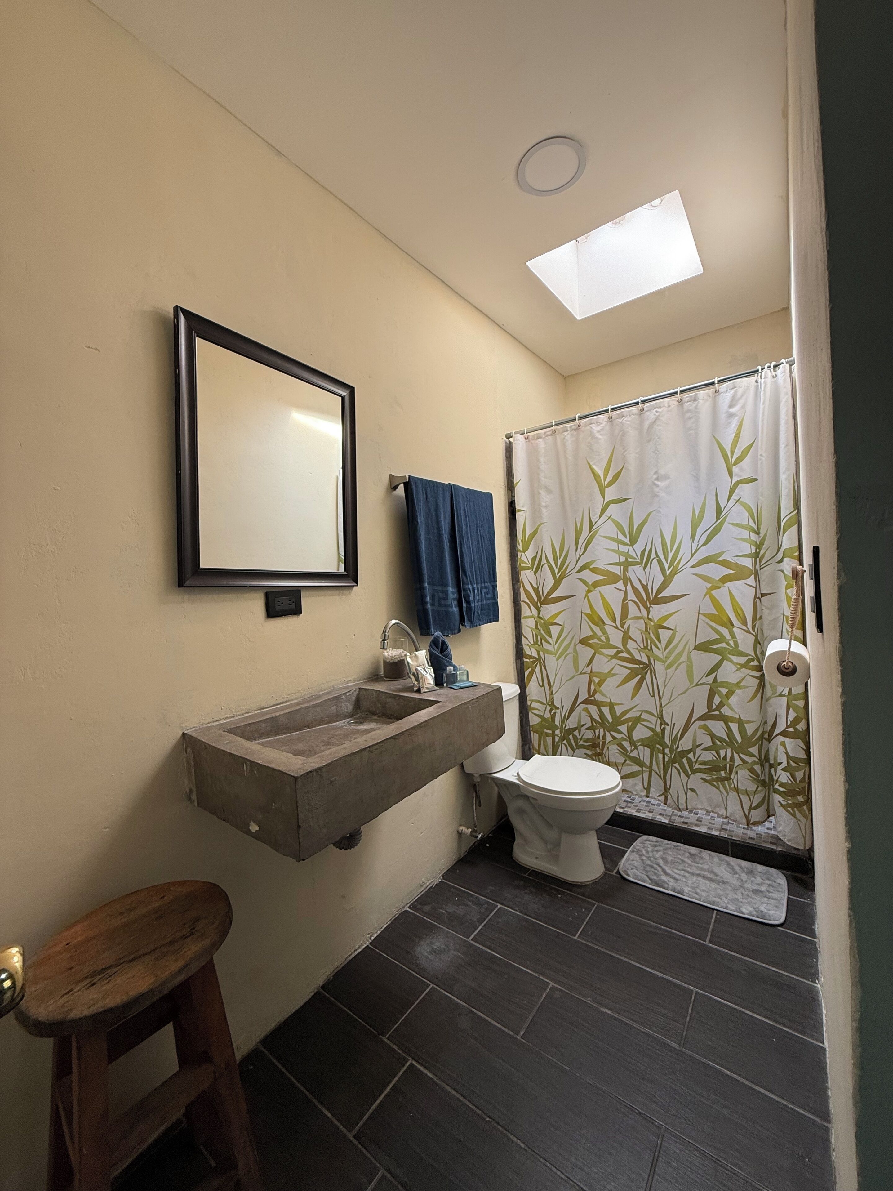 Un baño con lavamanos integrado, inodoro, espejo, taburete de madera, cortina de ducha de bambú y tragaluz.