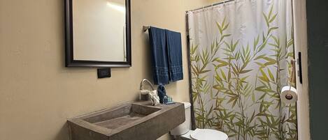A bathroom with a built-in sink, a toilet, a mirror, a wooden stool, a bamboo shower curtain, and a skylight.