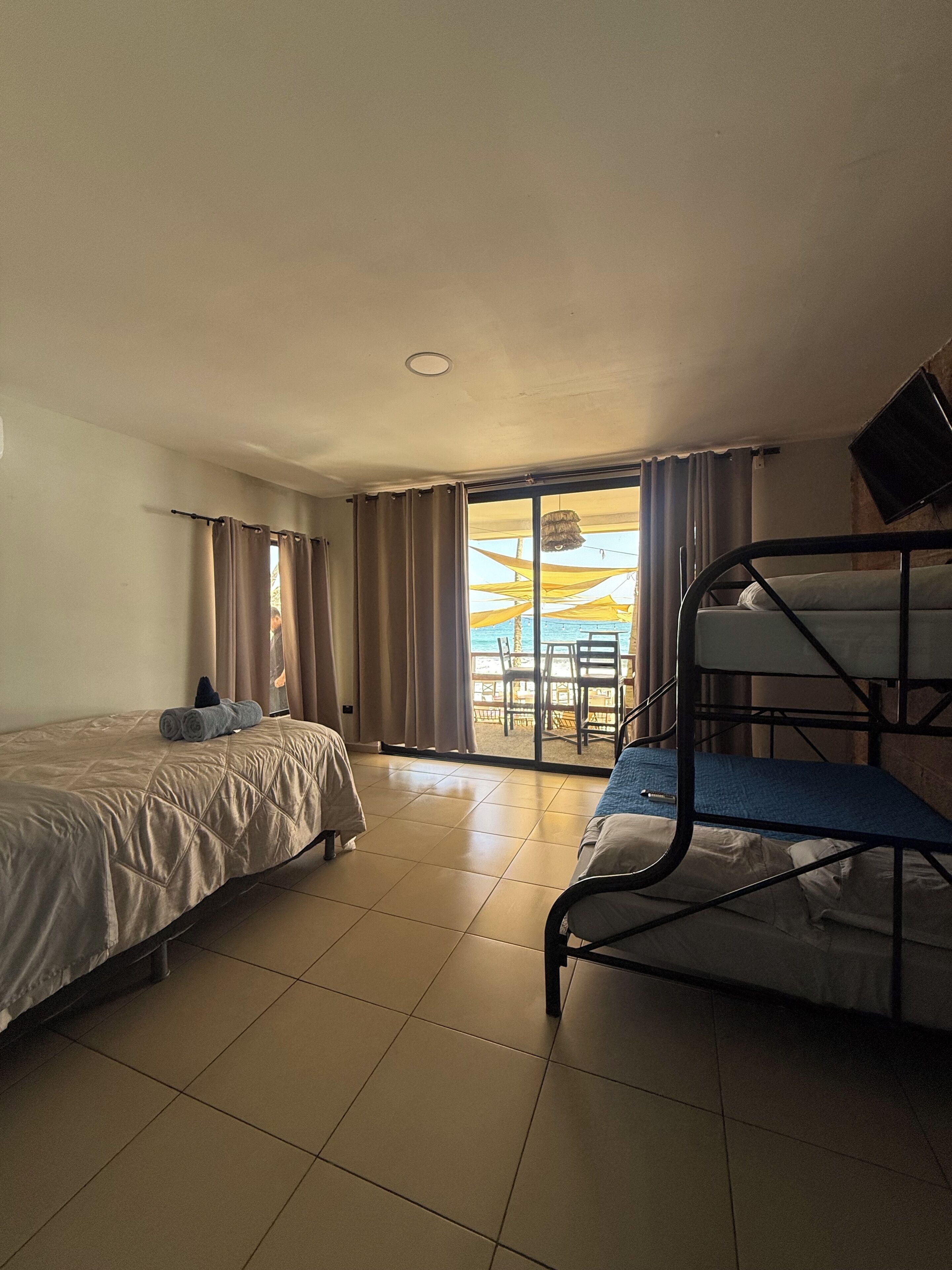 Quarto de hotel com duas camas, varanda com mesa e cadeiras, e vista para a praia.