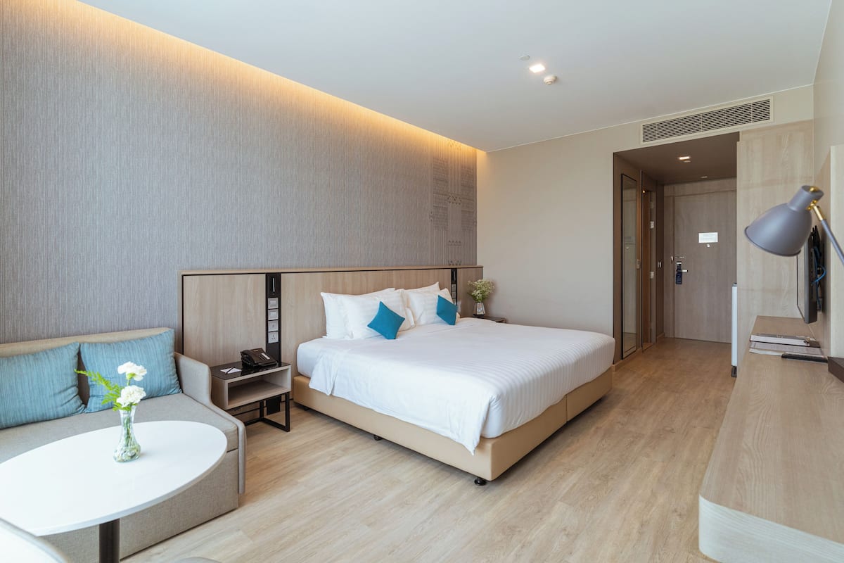 A modern hotel room with a large bed, a sofa, a small table with a vase, and a desk with a lamp.