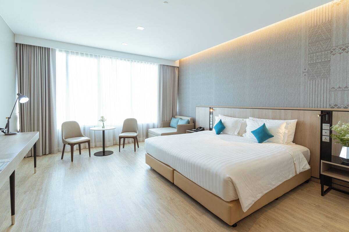 A modern hotel room with a large bed, a desk, two chairs, a small table, and a large window with curtains.