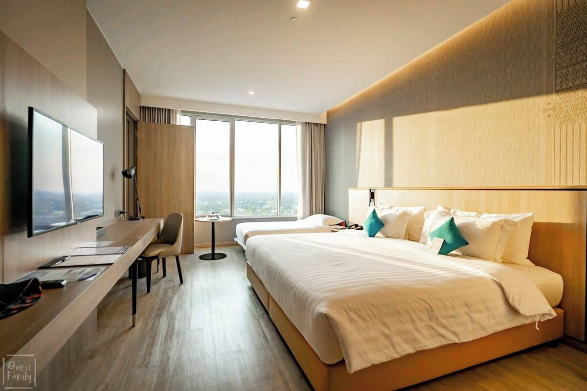 A hotel room with a large bed, a desk with a chair, a television, and a view of the city through large windows.