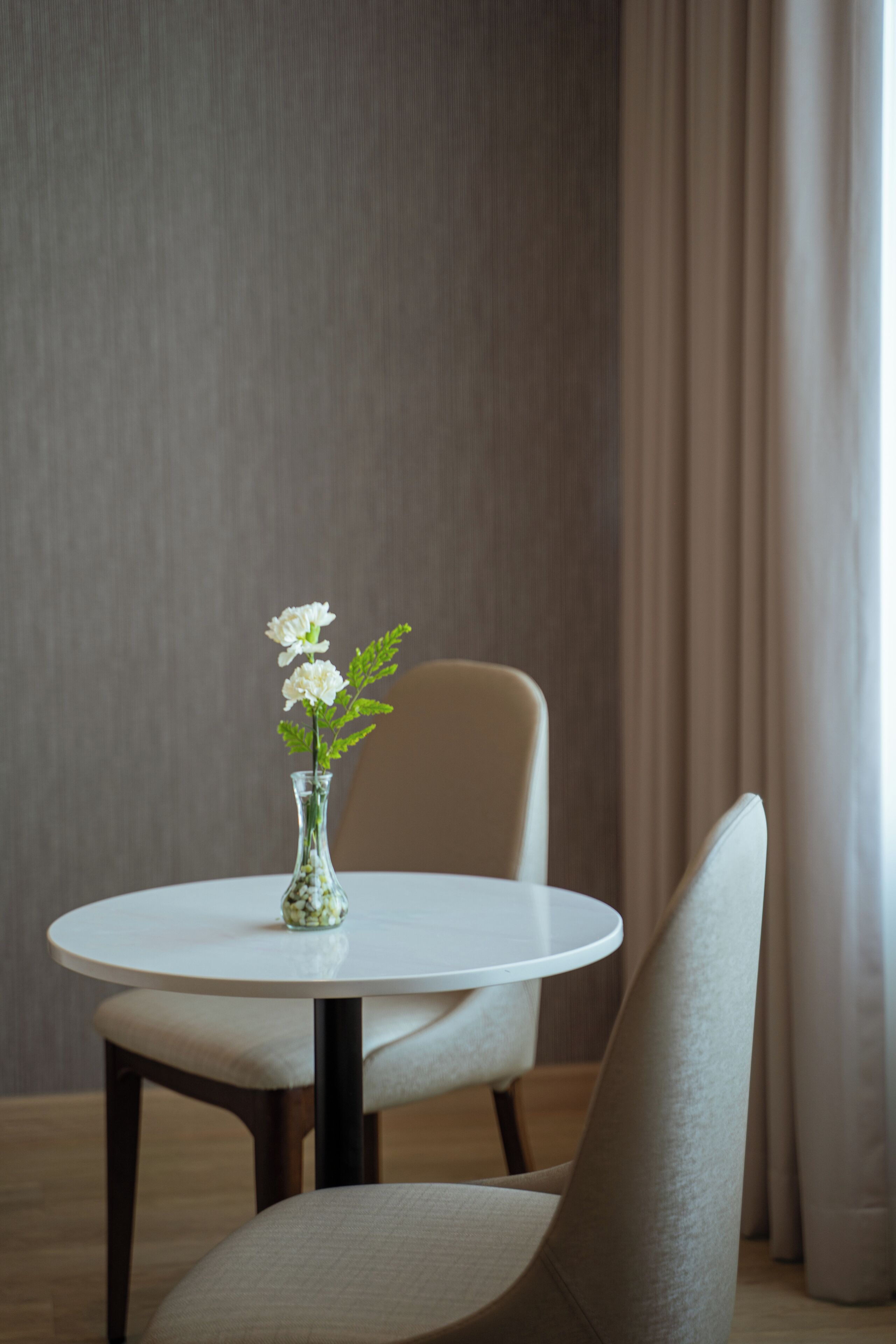 A round table with a vase of white flowers, a chair, and a curtain in the background.