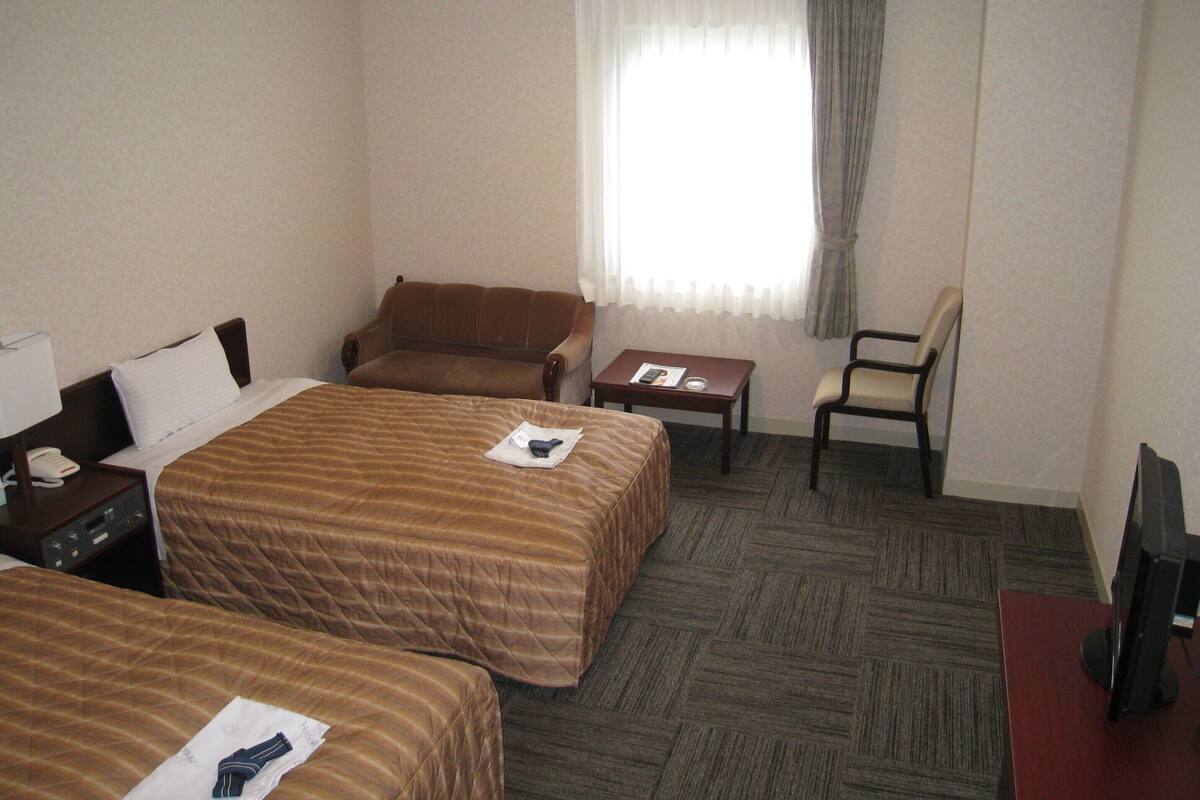 A hotel room with two beds, a desk, a chair, a TV, and a window with curtains.