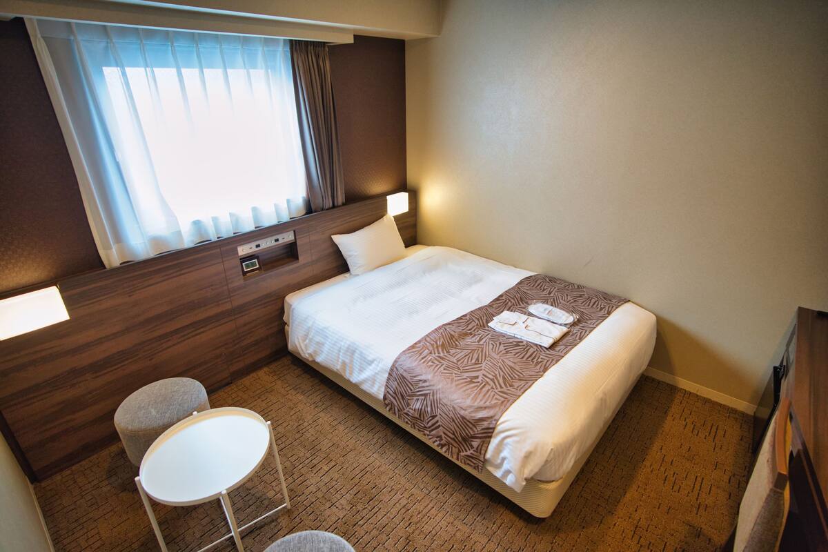 Ein Hotelzimmer mit einem Bett, einem kleinen Tisch, einem Stuhl, einem Nachttisch und einem Fenster mit Vorhängen.