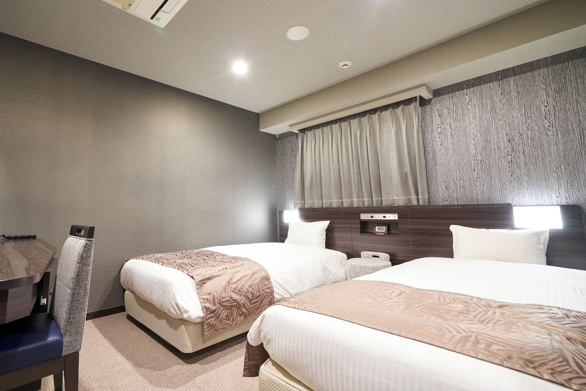 Ein Hotelzimmer mit zwei Betten, einem Schreibtisch, einem Sessel, einem Nachttisch und einem fenster mit Vorhängen.