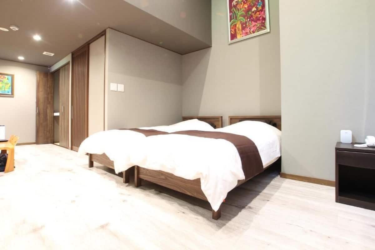 A hotel room with two beds, a wooden floor, a small desk with a chair, and a colorful painting on the wall.