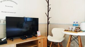 Une pièce avec un meuble TV en bois, un téléviseur à écran plat, une chaise blanche, une petite table avec des collations et des boissons, et une branche décorative accrochée au mur.