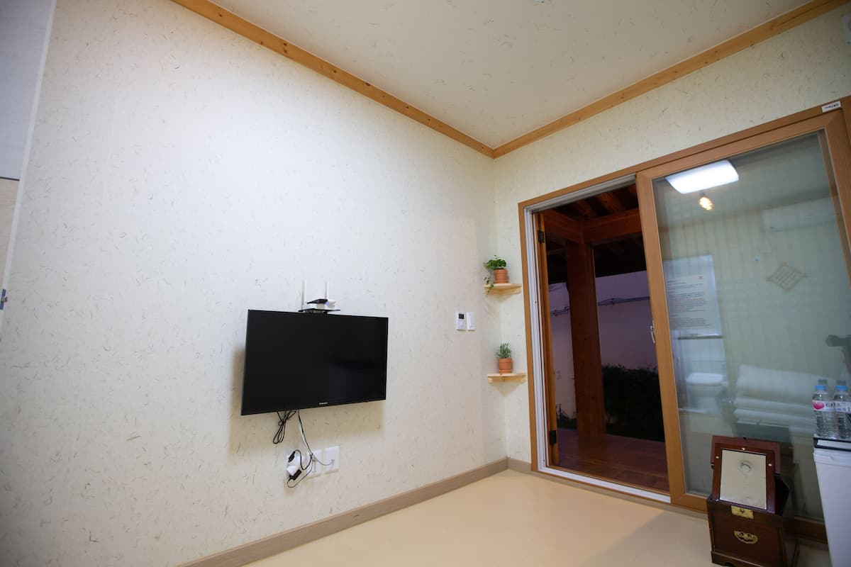 A room with a mounted TV, wooden beams on the ceiling, a sliding glass door, and a small shelf with a plant.