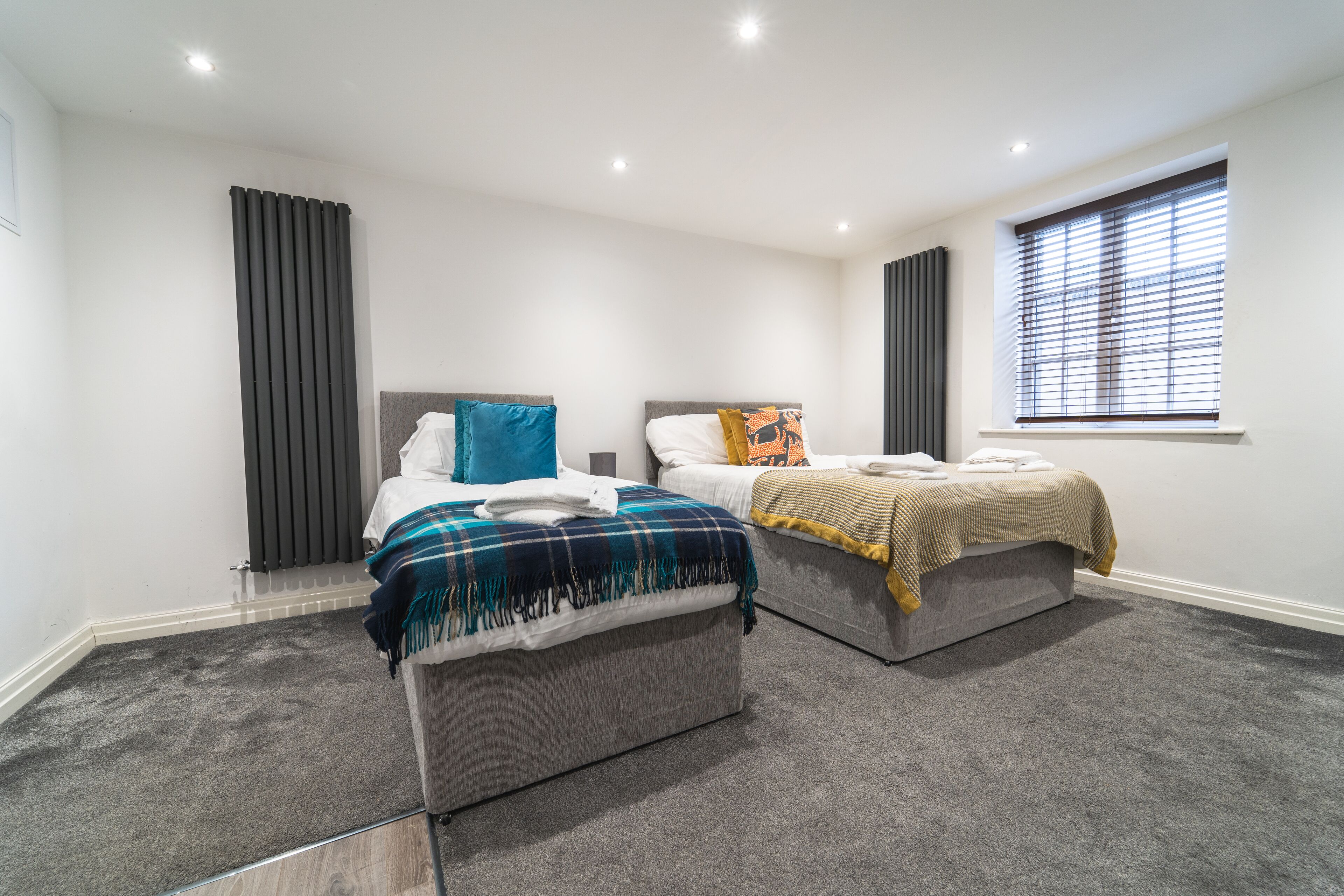 A modern bedroom with two beds, a large window with blinds, and a carpeted floor.