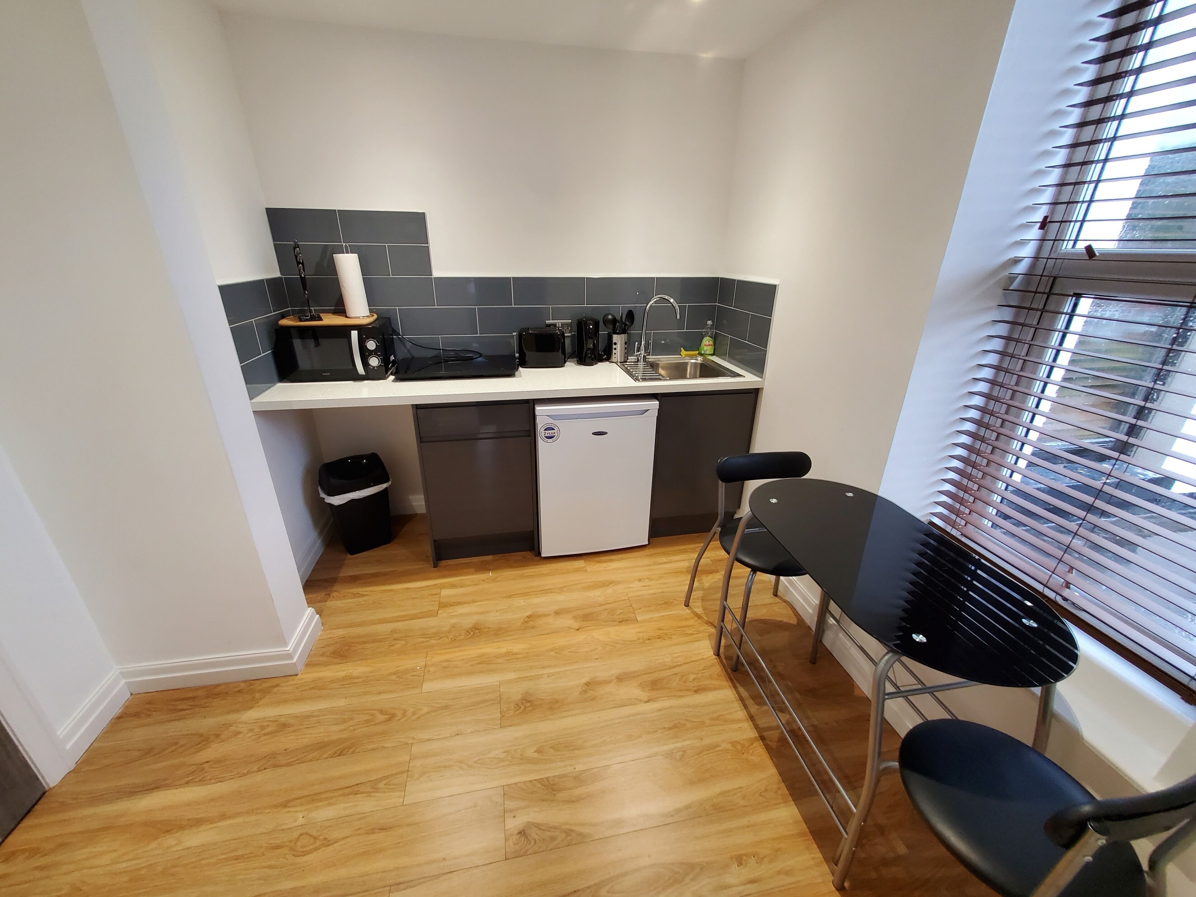 A compact kitchen with a dishwasher, a small refrigerator, a sink, and a window with blinds.