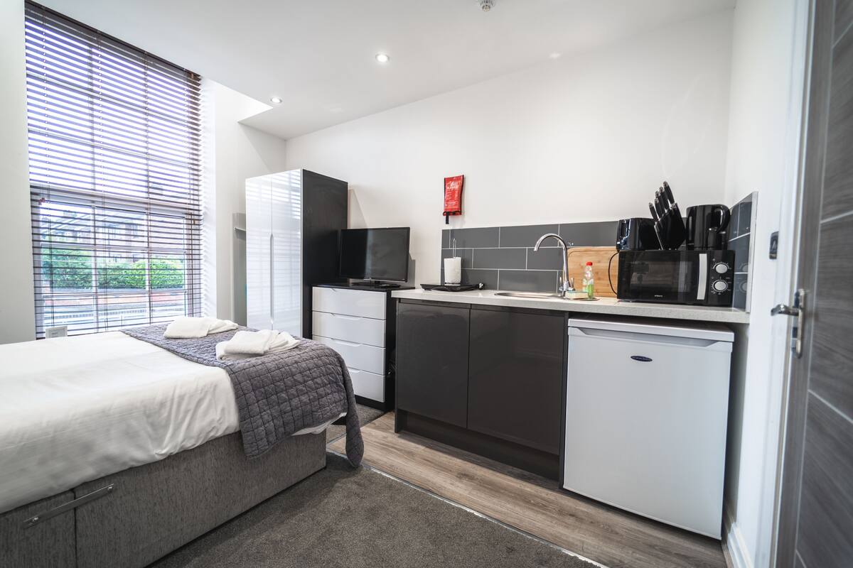 A modern hotel room with a bed, a small kitchenette, a TV, and a window with blinds.