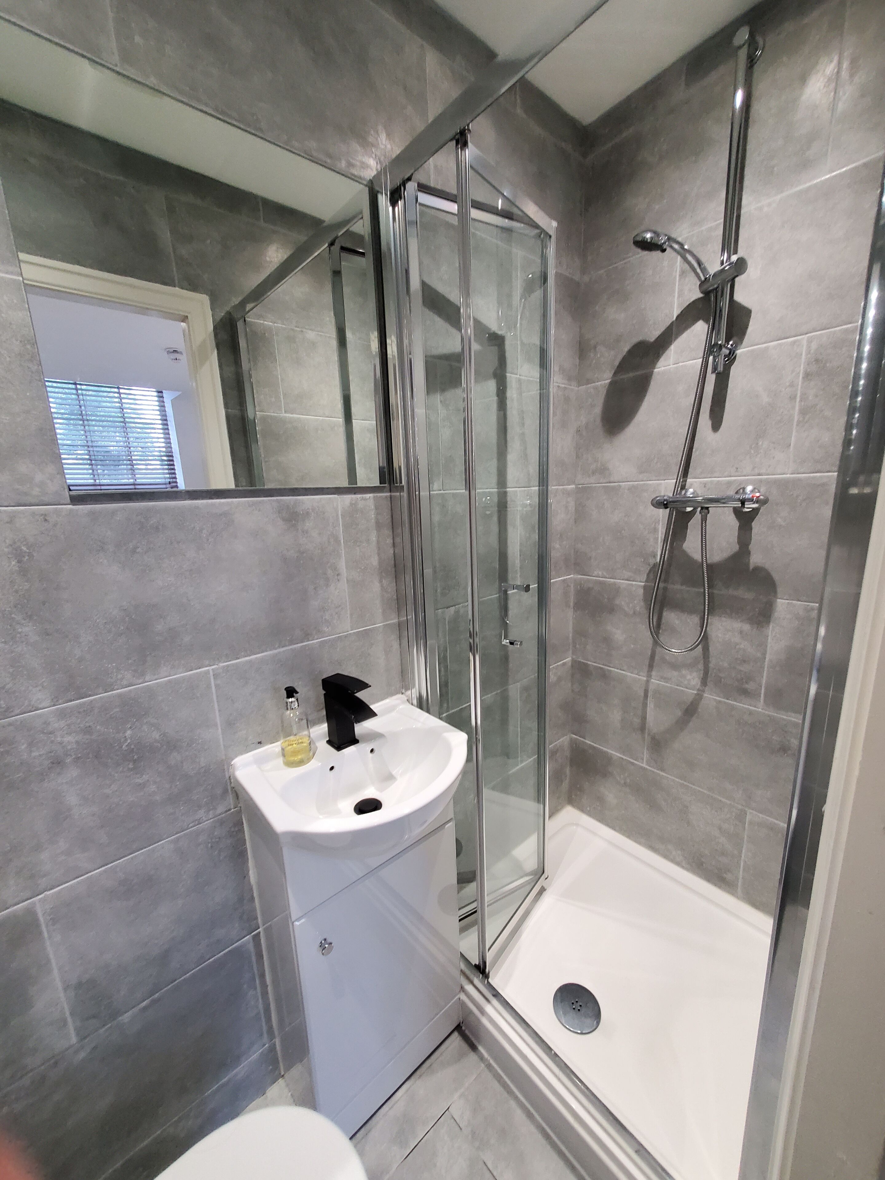 Un baño moderno con ducha envuelta en vidrio, un lavabo blanco y azulejos grises.