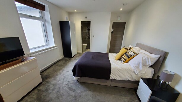A bedroom with a bed, a TV on a dresser, a window, and a door leading to a bathroom. - The Ashcroft Apartments (Manchester)