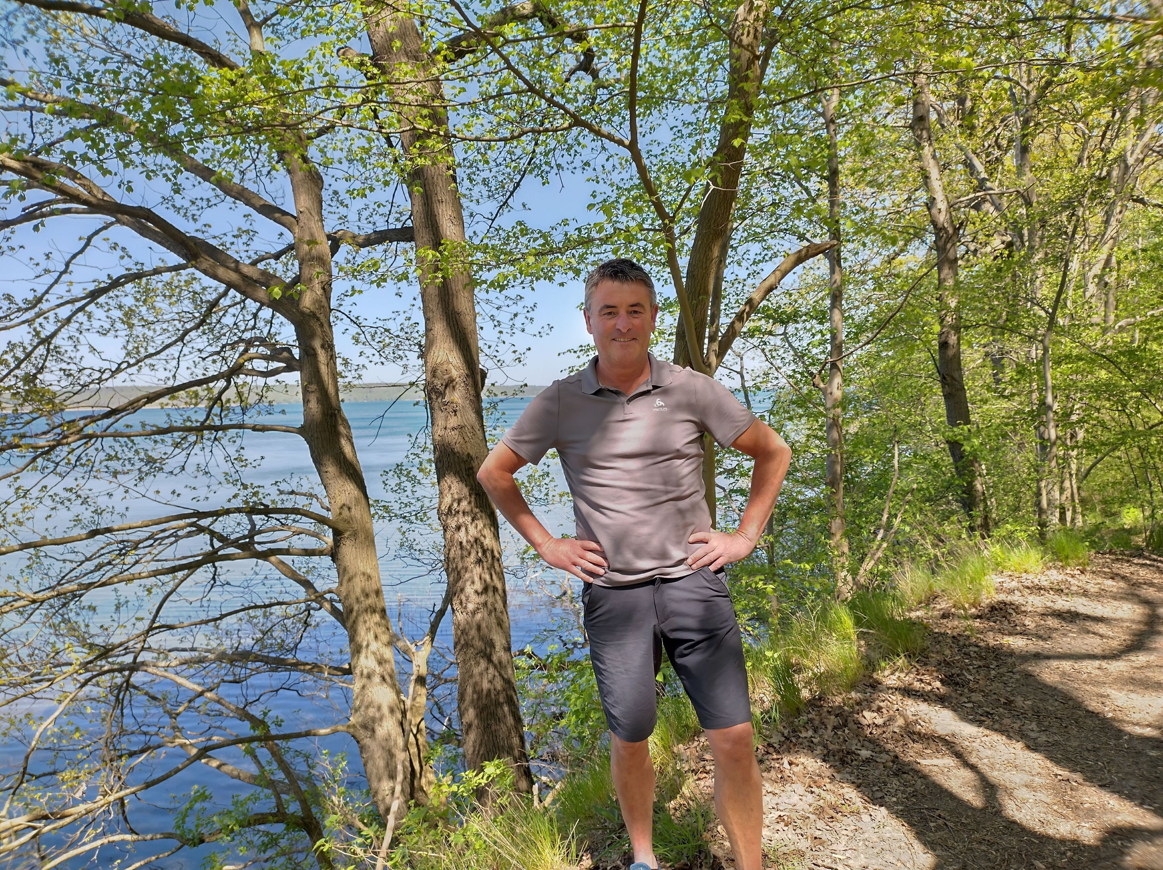 Une personne se tient sur un chemin de terre, entourée d’arbres et bordée par une étendue d’eau.