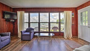 Una habitación con paredes de madera, una mesa de vidrio, dos sillas, un sofá, un televisor montado en la pared y ventanas grandes con cortinas.
