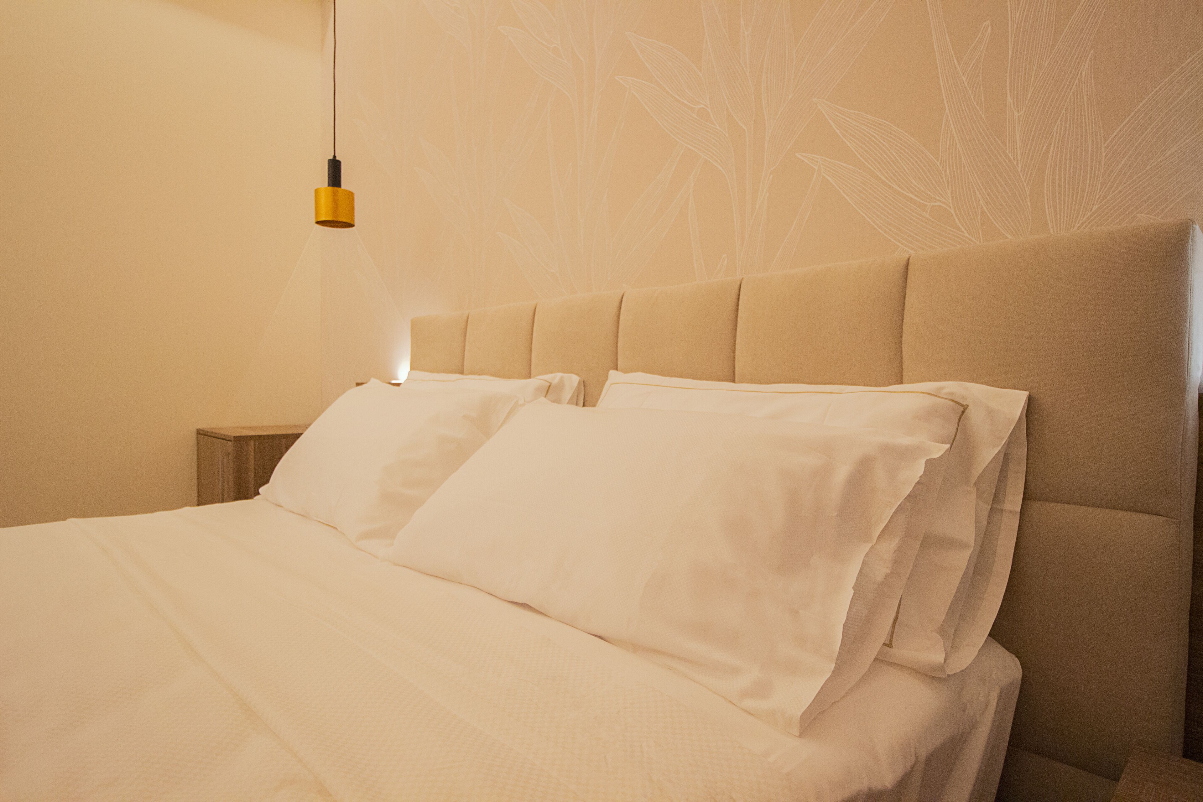Una cama bien hecha con sábanas blancas y un cabecero, una mesita de noche y una pared con un discreto estampado de hojas.