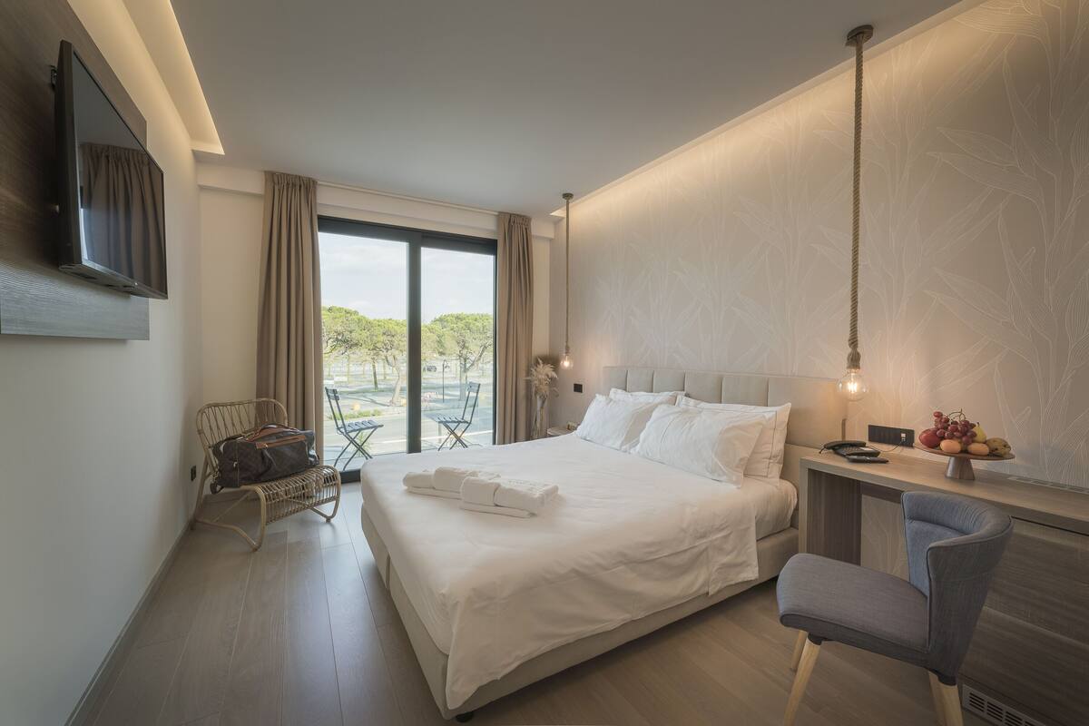 Una habitación de hotel moderna con una cama grande, un escritorio con silla, un televisor en la pared y vistas al exterior.