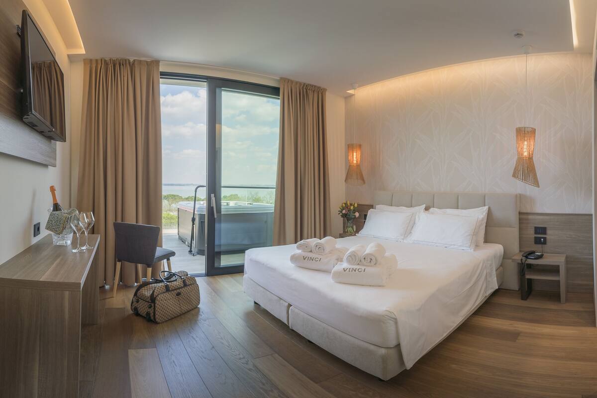 Habitación de hotel con una cama grande, un escritorio, una silla, un televisor y un balcón con cortinas.