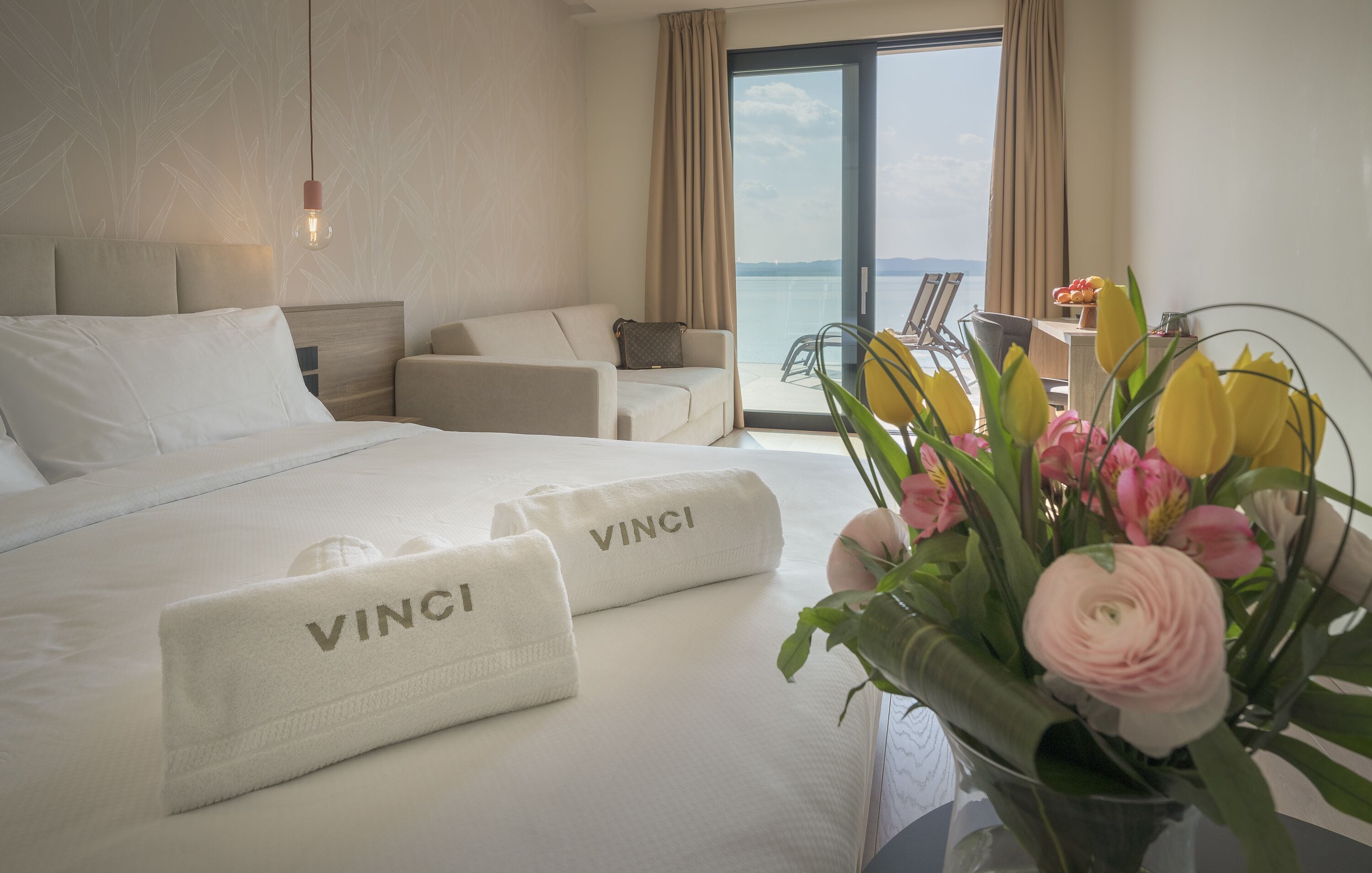 Habitación de hotel con cama, dos toallas VINC1, un florero con flores y vista al mar por la ventana.