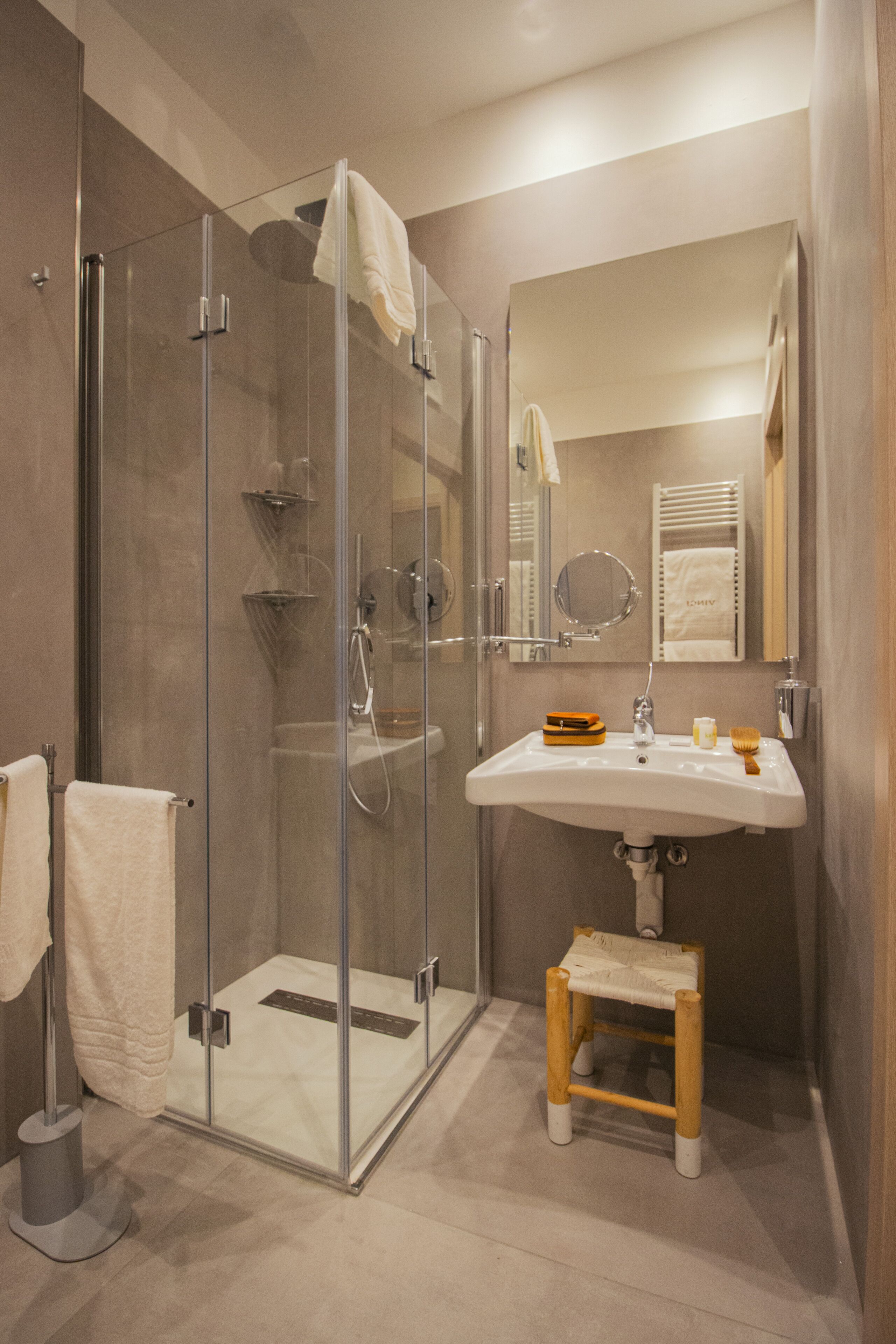 Un baño moderno con ducha envuelta en vidrio, un lavabo con artículos de aseo y un taburete de madera.
