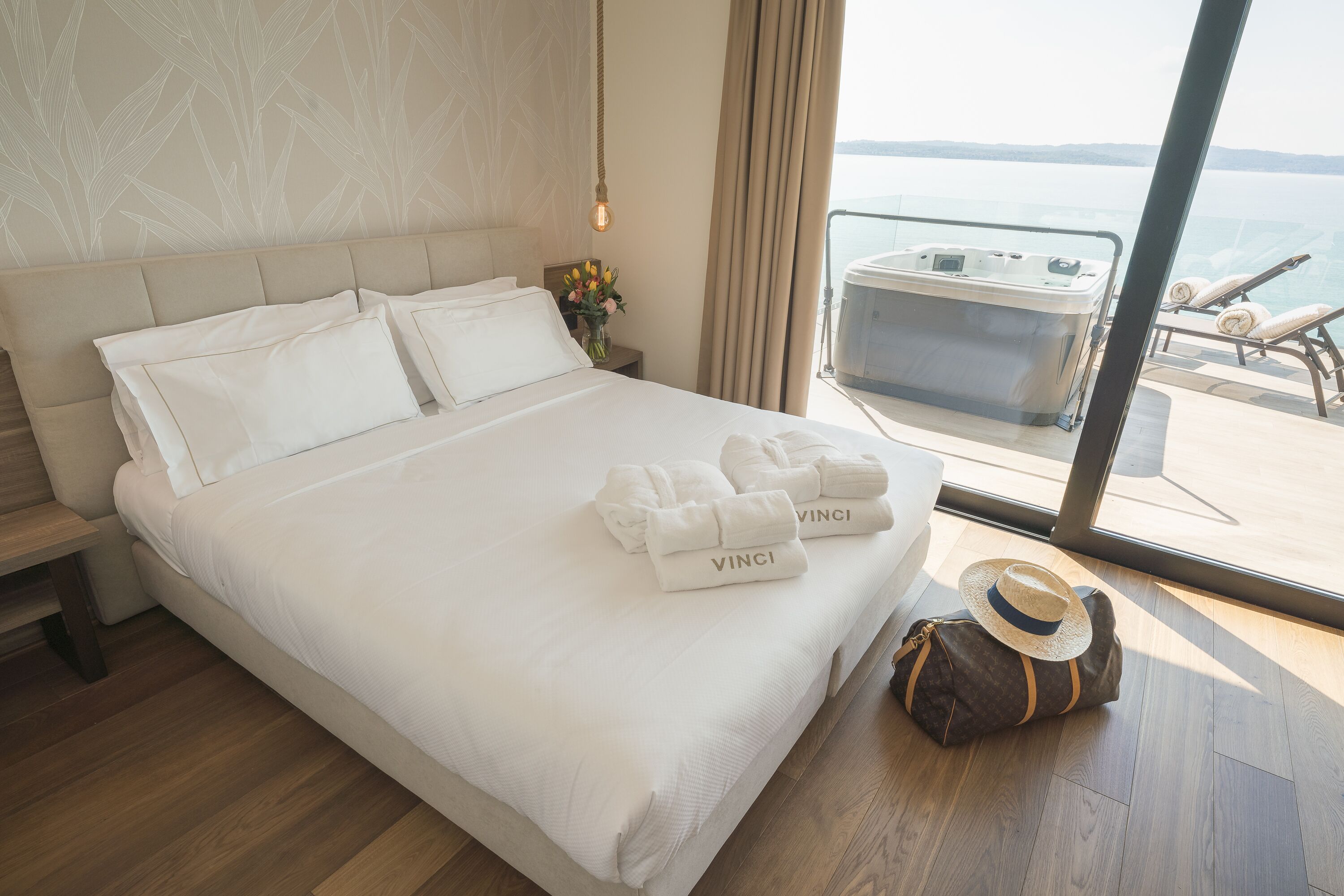 A hotel room with a large bed, a view of the sea, a hot tub, and a suitcase on the floor.