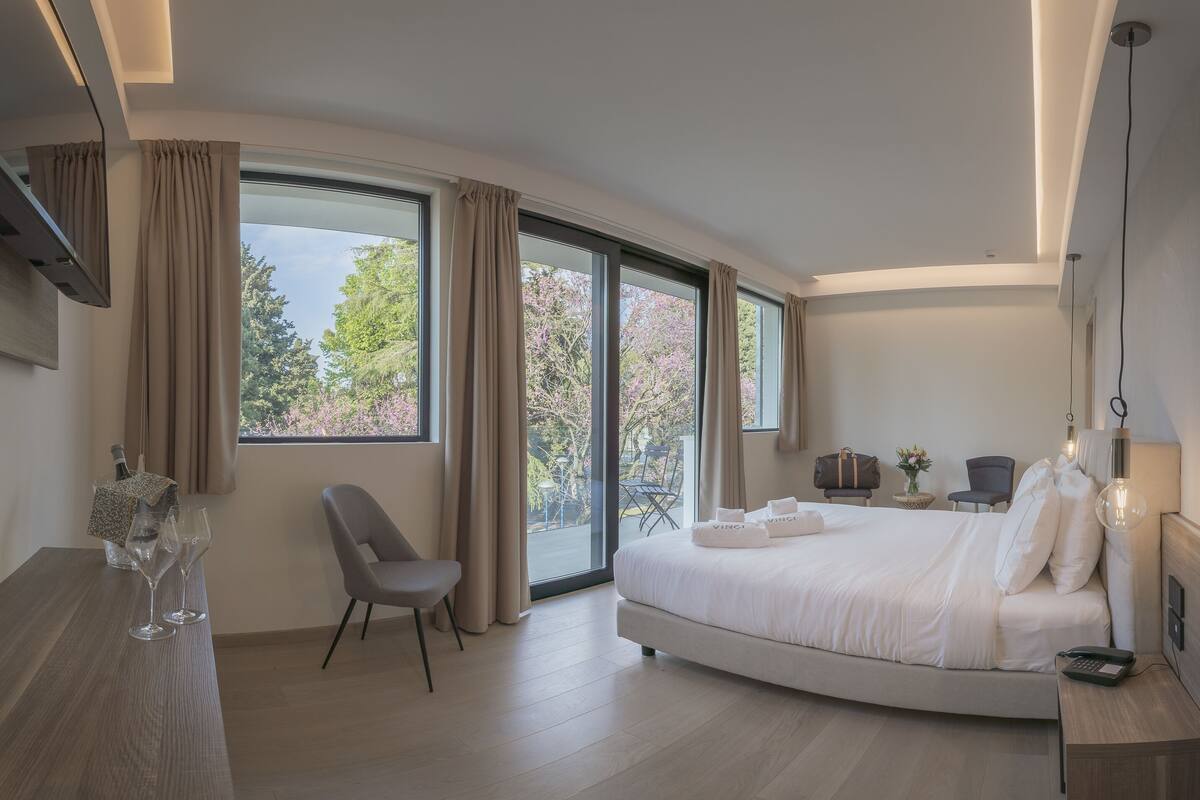 Habitación de hotel moderna con una cama curvada, ventanas amplias, zona de comedor y vistas a árboles.