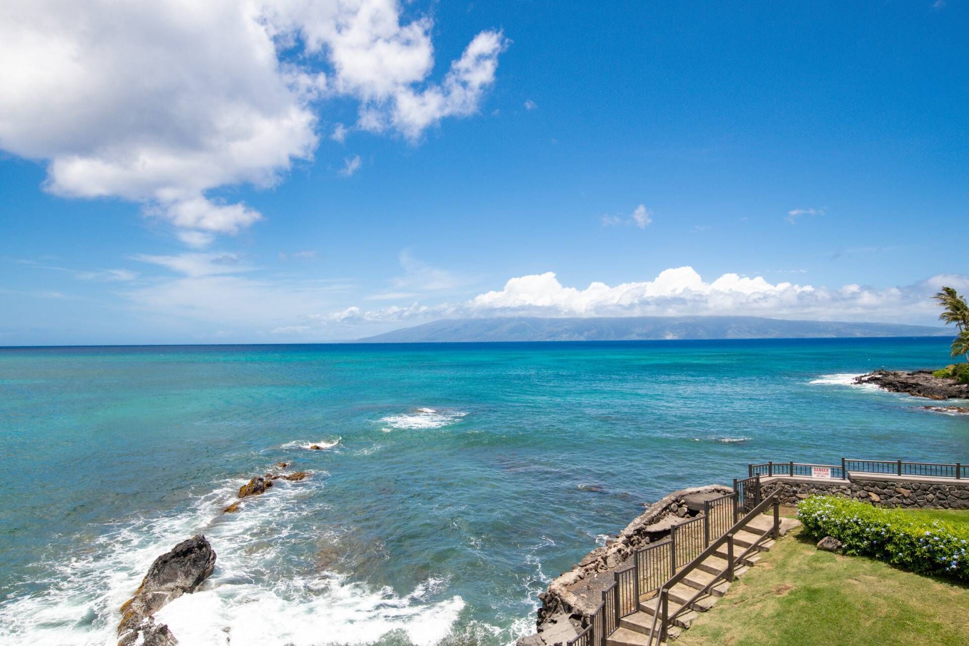 A coastal view with clear skies, turquoise waters, rocky shoreline, and a wooden staircase leading to a viewing platform.