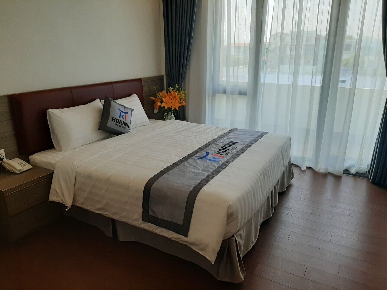 Una camera d'albergo con un letto, un comodino, un telefono, un vaso con fiori e vista all'esterno attraverso tende leggere.