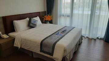 Una camera d'albergo con un letto, un comodino, un telefono, un vaso con fiori e vista all'esterno attraverso tende leggere.