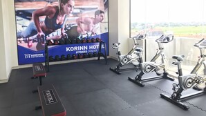 Une salle de sport équipée de matériel d’exercice, d’un grand écran de télévision diffusant des images sur le thème du fitness, et offrant une vue sur l’extérieur grâce à de grandes fenêtres.