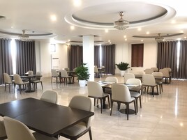 Uma sala de conferências ampla, com várias mesas e cadeiras, um candelabro e grandes janelas com cortinas.