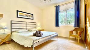 A bedroom with a bed, a chair, a window with curtains, a chandelier, and a painting on the wall. - Mila 3 bedrooms, air-con, free wifi (Ametlla de Mar)