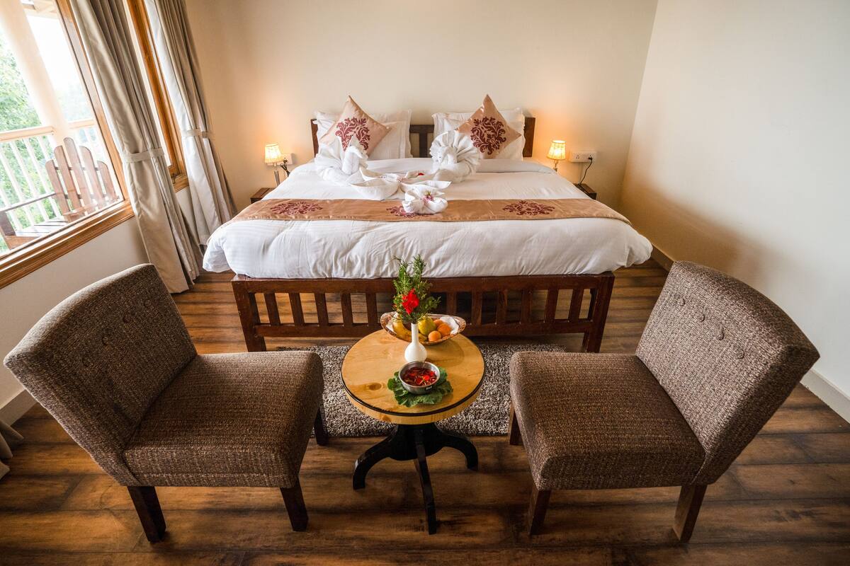 A neatly arranged hotel room with a large bed, two armchairs, a small round table with a vase of flowers, and a view of the outdoors through a window.
