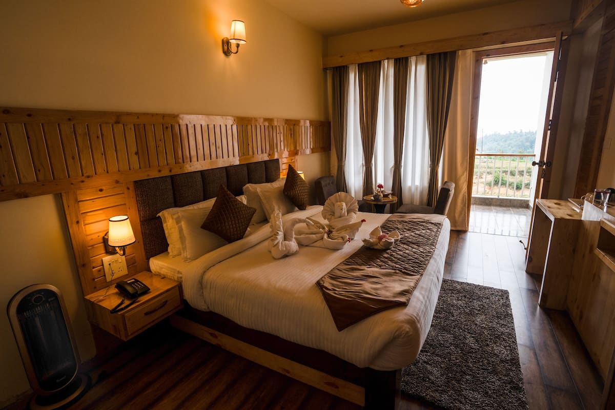 A hotel room with a large bed, wooden headboard, bedside tables with lamps, a small table with a vase, and a view of the outdoors through a door.