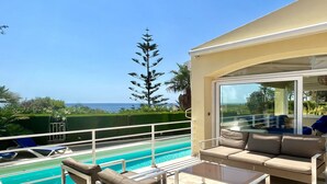 A poolside area with a sofa and chairs, a glass table, and a view of the ocean.
