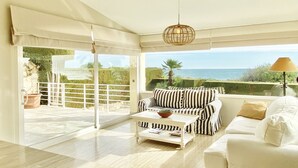 A spacious living room with a white sofa, striped armchair, and a coffee table. Large glass doors lead to a balcony with a view of the sea.