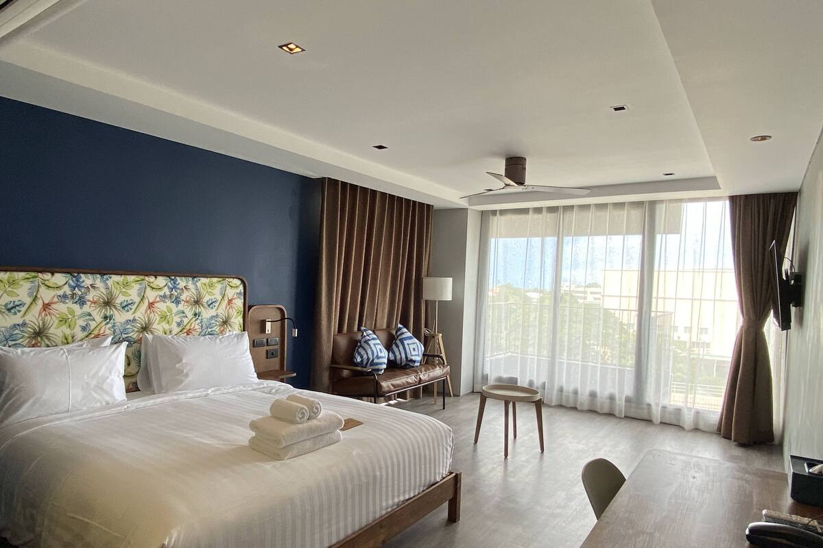 A hotel room with a large bed, a desk, a chair, a window with curtains, and a ceiling fan.