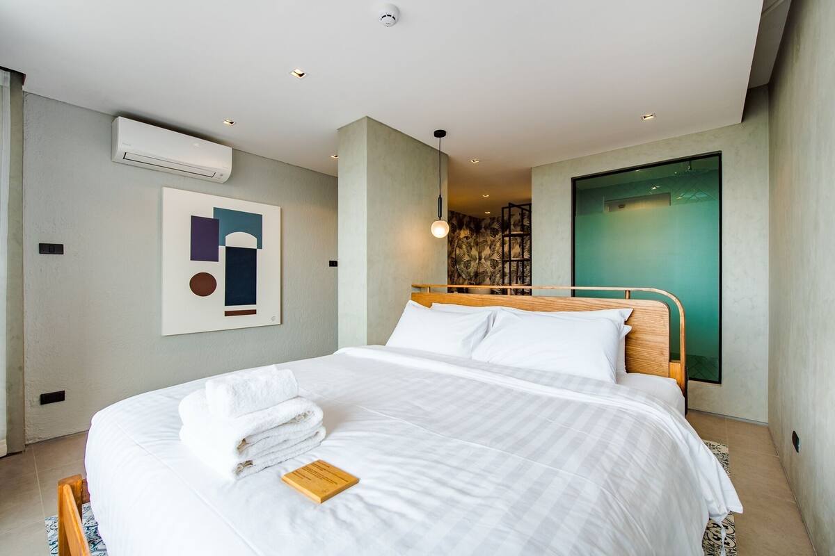 A modern hotel room with a neatly made bed, a large abstract painting on the wall, and a view of the city through a large window.