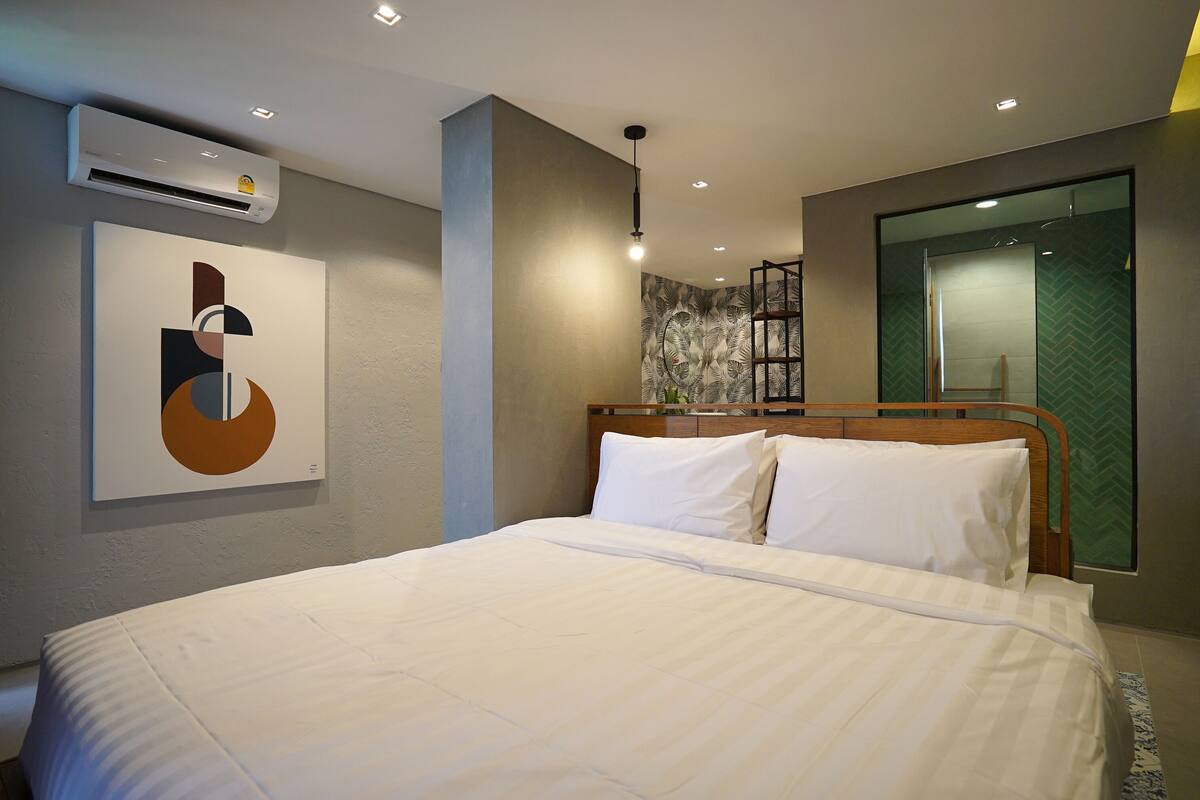 A modern hotel room with a large bed, a wall-mounted air conditioner, a geometric artwork, a hanging light fixture, and a green patterned wall.